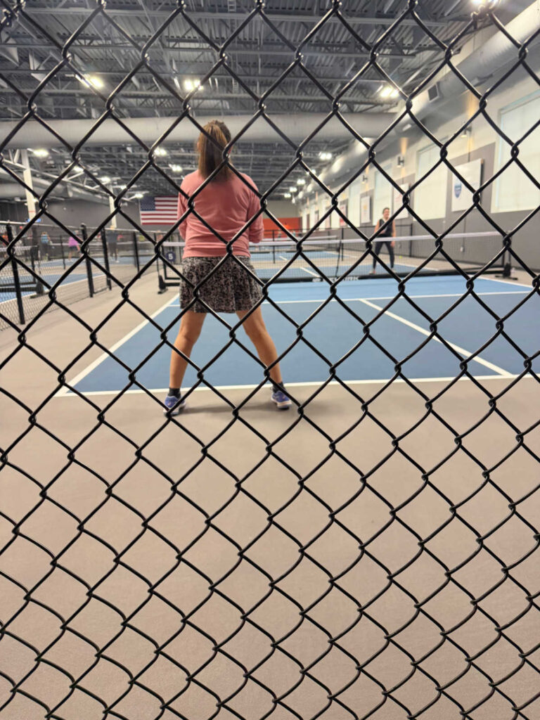 A person stands ready on an indoor pickleball court, viewed through a chain-link fence. The court, framed by industrial lighting and walls, is alive with anticipation. An opponent waits across the net, embodying the competitive spirit measured by DUPR in pickleball.