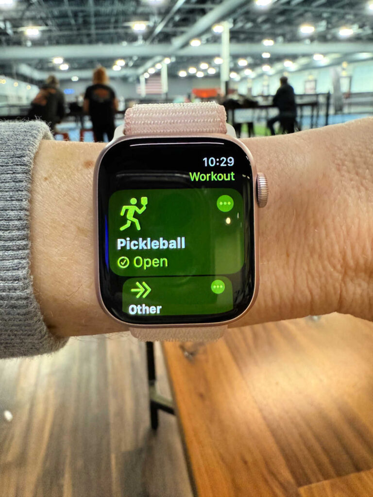 A person wears a smartwatch displaying a Pickleball workout option marked as Open, highlighting the health benefits of Pickleball. The background reveals an indoor setting with people sitting at tables.