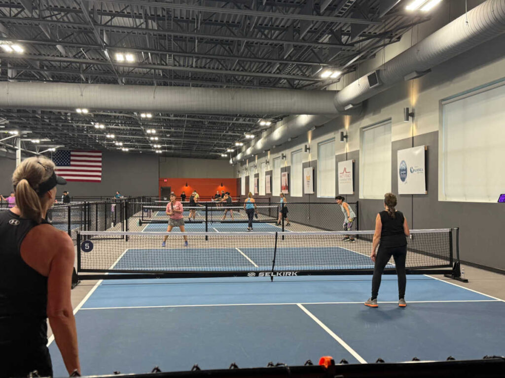 A group of people enjoying some indoor pickleball action, dinking and smashing on several courts. Players wield their paddles with flair, hitting the ball under high ceilings. The facility showcases an American flag and logo banners, adding a vibrant touch to the pickleball scene.