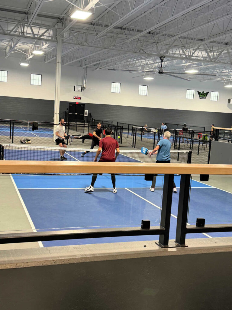 Four people are playing pickleball indoors on a blue court, engaged in a lively doubles match. The facility boasts bright lighting and a high ceiling, perfect for executing what is rally scoring in pickleball with precision as the game intensifies on each side of the net.