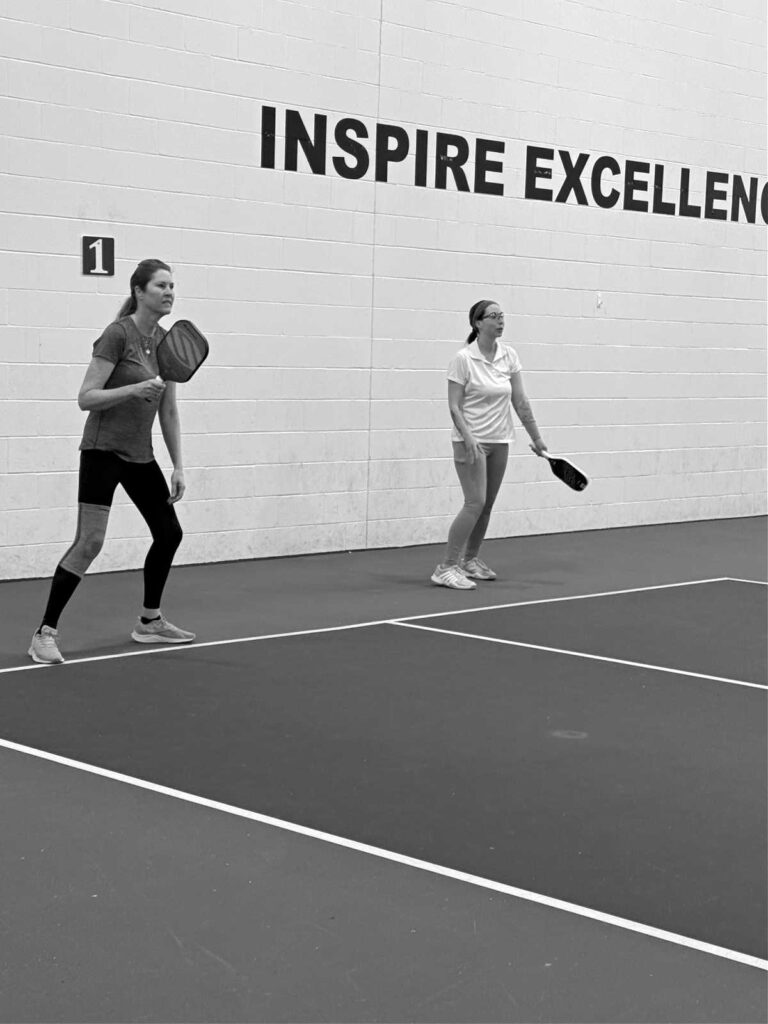 Two women stand ready on an indoor court, each holding a paddle, eager to say "Let's Play Pickleball." A motivational phrase INSPIRE EXCELLENCE is partially visible on the wall behind them. The scene is captured in striking black and white.