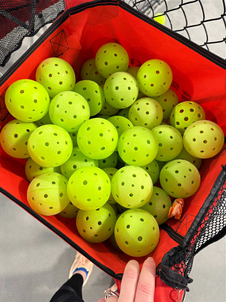 A person holding a red basket filled with numerous yellow pickleballs, each featuring multiple holes. The basket, partially covered by black mesh, prompts curiosity about what is the difference between indoor and outdoor pickleball balls.