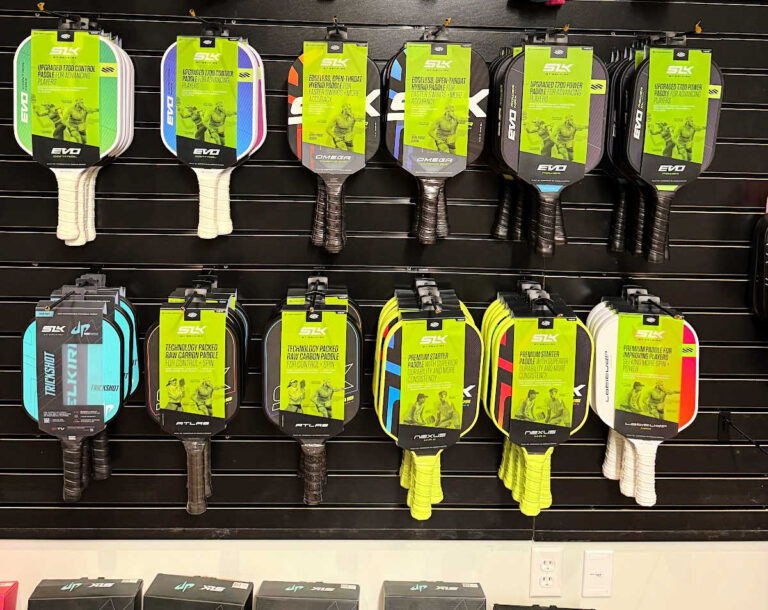 A display of various pickleball paddles hangs on a black slat wall in a sports store, showcasing an array of designs and colors. This selection of pickleball equipment includes paddles with green and black packaging, making it a vibrant focal point for enthusiasts.