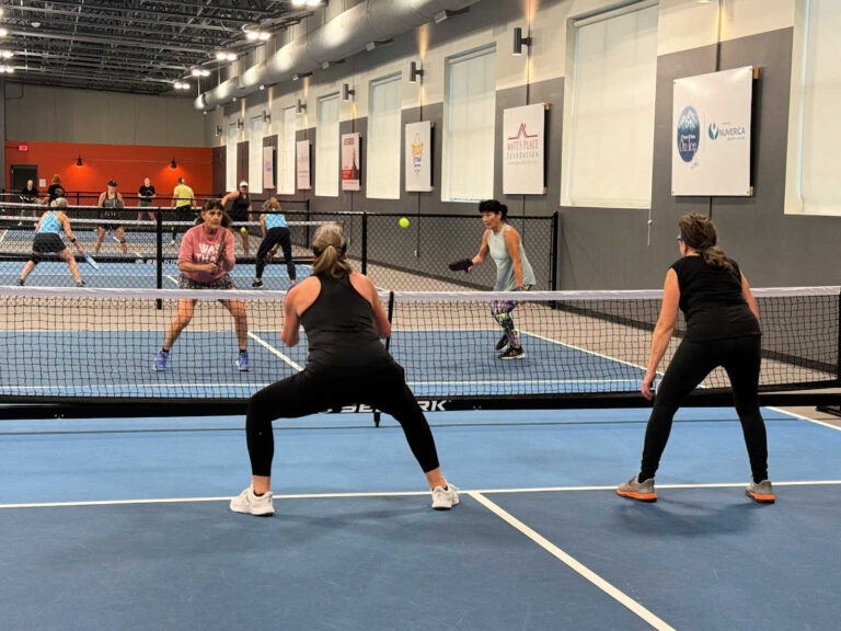 People playing pickleball indoors on blue courts. Two women are in the foreground, facing two opponents. The facility, perfect for showcasing what pickleball is all about, has high ceilings and multiple courts visible in the background.