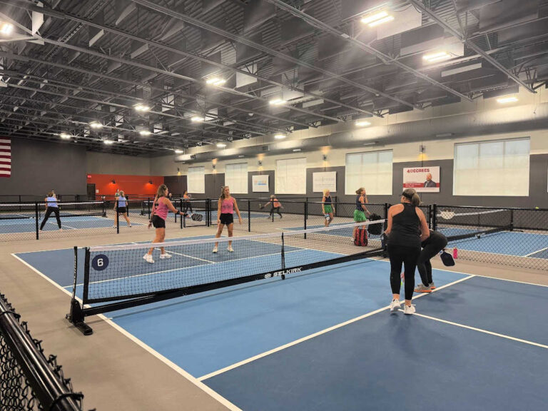 A group of people playing pickleball on indoor courts showcases what pickleball is all about. Bright lighting illuminates multiple courts as players engage in various stages of the game. An American flag hangs on the wall above gray banners and windows, adding to the vibrant atmosphere.