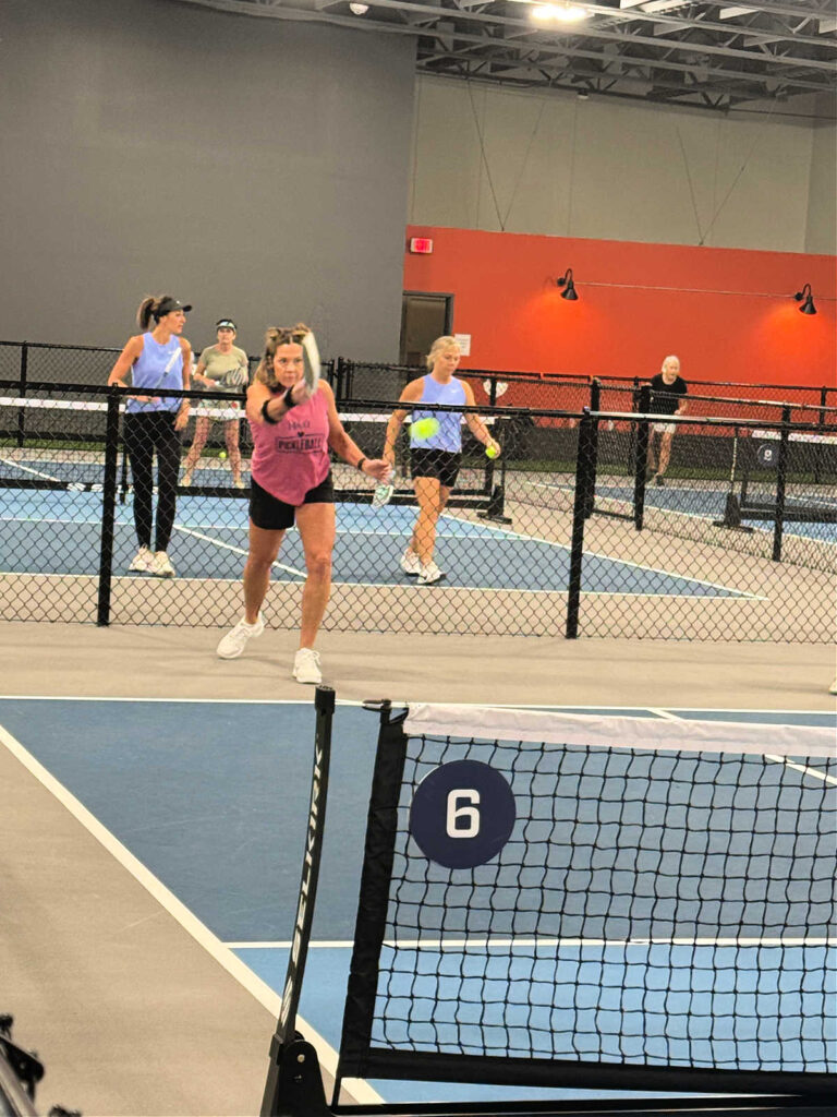 A group of people playing pickleball indoors on blue courts, where the starting score of a doubles game is 0-0-2. A woman in a pink shirt stands at the net, preparing to hit the ball, while others watch from behind the fence. The court is marked with the number 6.