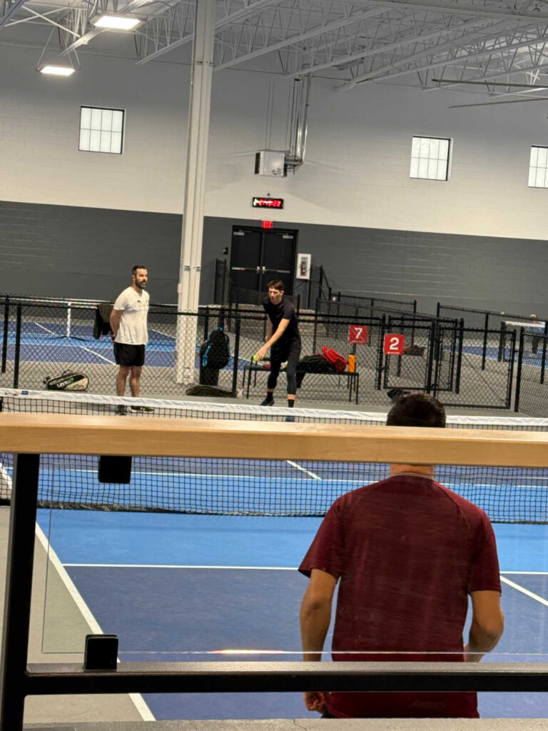 In an indoor setting that almost resembles a Pickleball Open Play, three players are engaged on blue tennis courts. One is poised to serve; another stands ready, while a third observer watches from the foreground. A black fence encloses the action-packed scene.