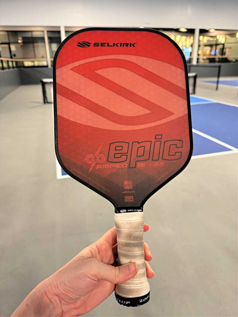 A hand holding a red Selkirk Epic pickleball paddle, touted as a good pickleball paddle by enthusiasts, showcases its branding and design details against the backdrop of a blurred indoor court.