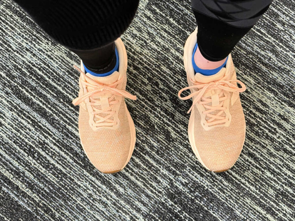 A person wearing peach-colored pickleball shoes stands on a textured, striped carpet. The shoes are paired with black leggings, and theres a hint of blue pickleball socks peeking above the sneakers.