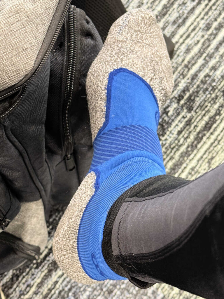 A foot wearing a stylish blue pickleball sock with gray slipper lies on a patterned carpet. Partially visible beside it is a black and gray bag, ready for adventures or perhaps to hold your favorite pickleball socks.