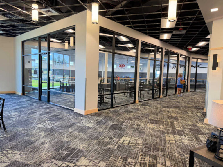 Indoor Pickleball Courts Post Falls, several people work at desks, while one person walks along the hallway by the glass walls.