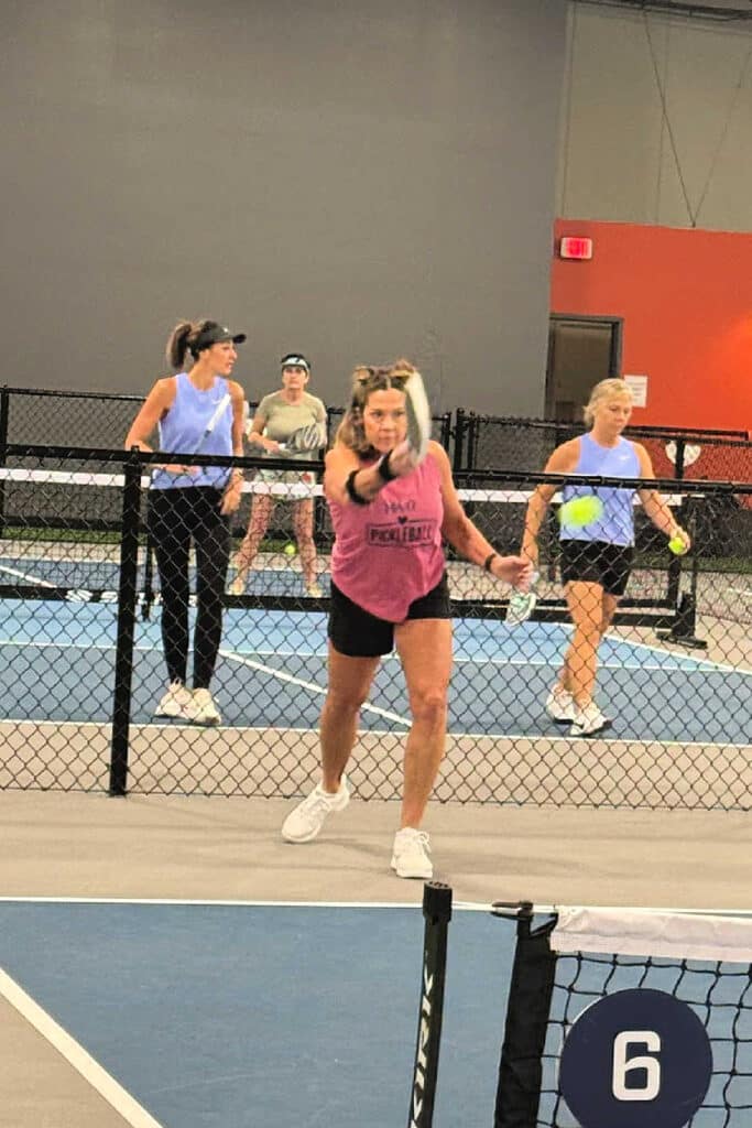 Four women play pickleball on an indoor court. One in a pink tank top swings her paddle, perhaps attempting a lob—an important shot when learning what is a lob in pickleball—while the others prepare and watch in this active, competitive scene.