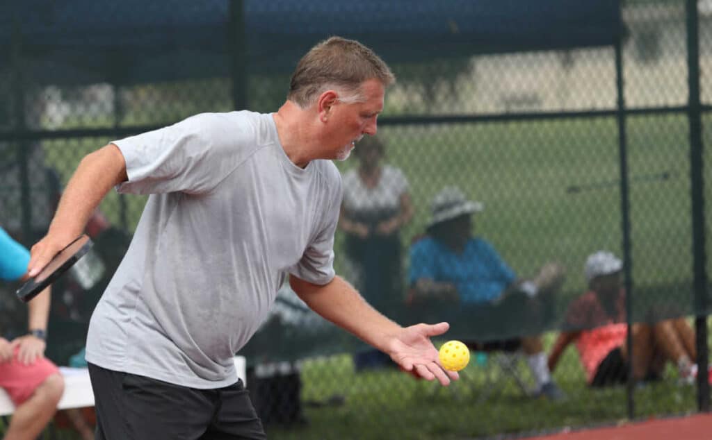 A man in a gray shirt prepares to serve during a pickleball game, carefully following pickleball serving rules as he holds a paddle and yellow ball on an outdoor court with a chain-link fence and spectators in the background.