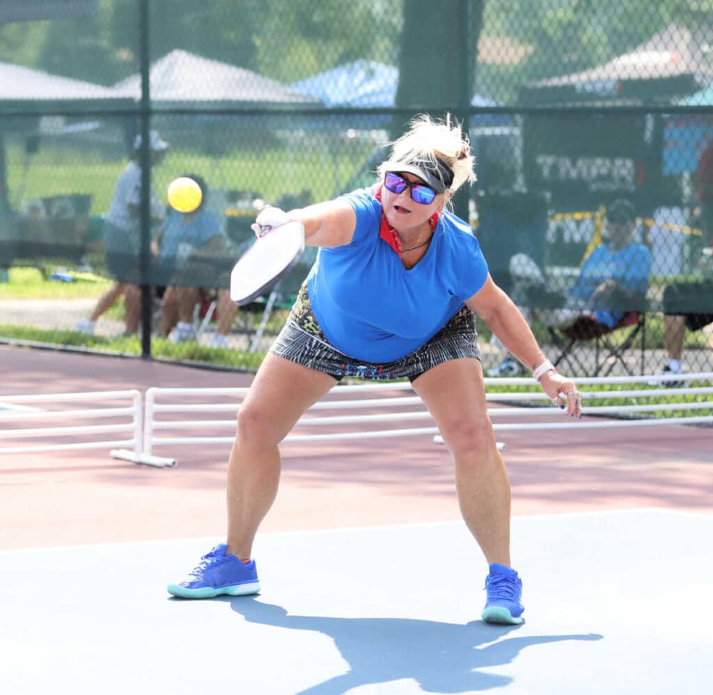 A woman in athletic clothing and sunglasses plays pickleball on an outdoor court, reaching forward with her paddle to hit the ball.