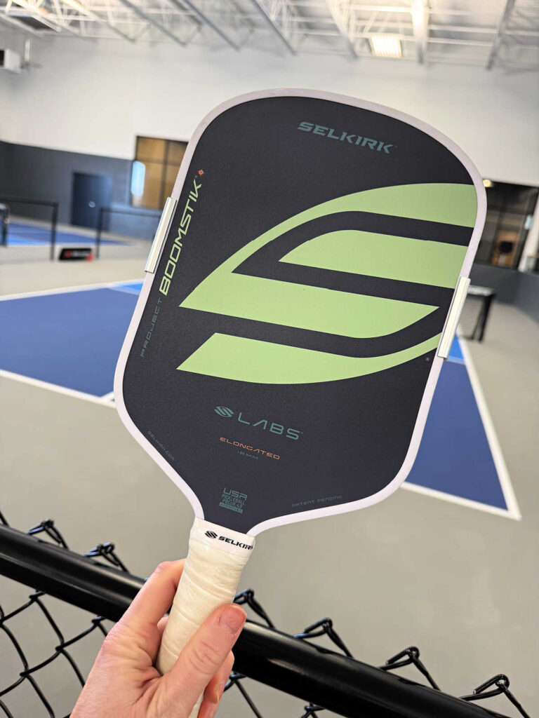 A hand holds a Selkirk Labs Boomslang 740SL pickleball paddle in front of a blue indoor pickleball court, with a black chain-link fence in the foreground.
