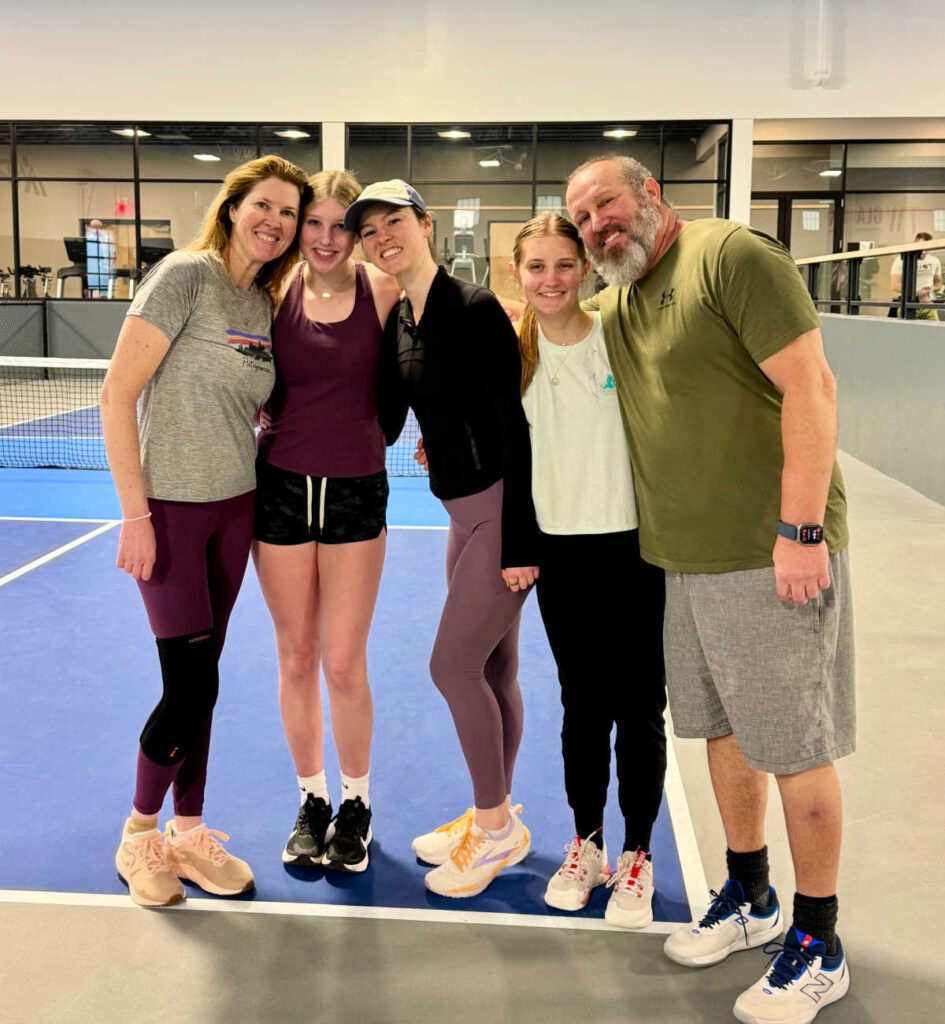 Five people stand smiling together on an indoor tennis or pickleball court, dressed in athletic wear. This about me moment captures four women on the left and one man on the right, all appearing happy and relaxed.