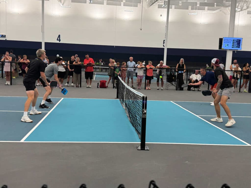 Four people play a doubles pickleball match indoors at The Flying Pickle Post Falls while a crowd watches from behind a fence. Players are poised near the net, focused on the game. The court has blue and gray flooring, and a screen displays the score.