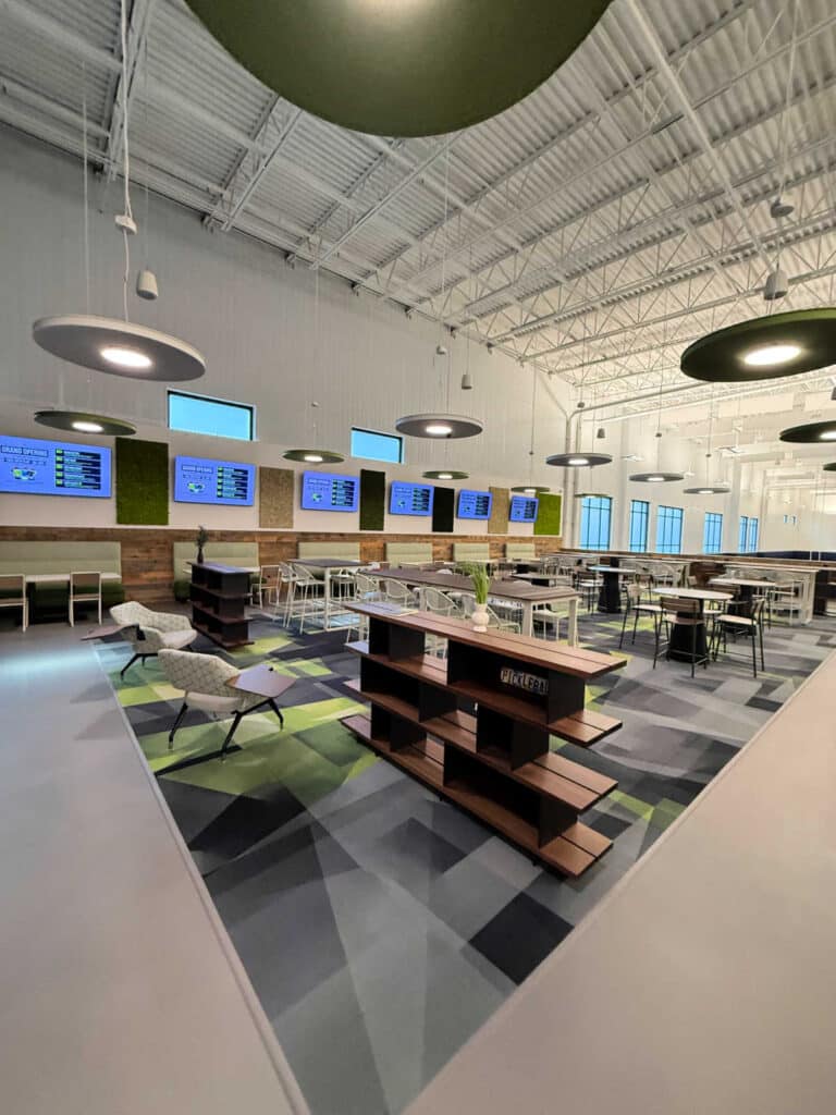 Modern cafeteria with green and gray decor, round ceiling lights, dining tables, chairs, lounge seating, digital menu screens on the wall, large windows, and a geometric patterned floor—The Flying Pickle Post Falls offers a clean and spacious environment for diners to relax and enjoy their meal.