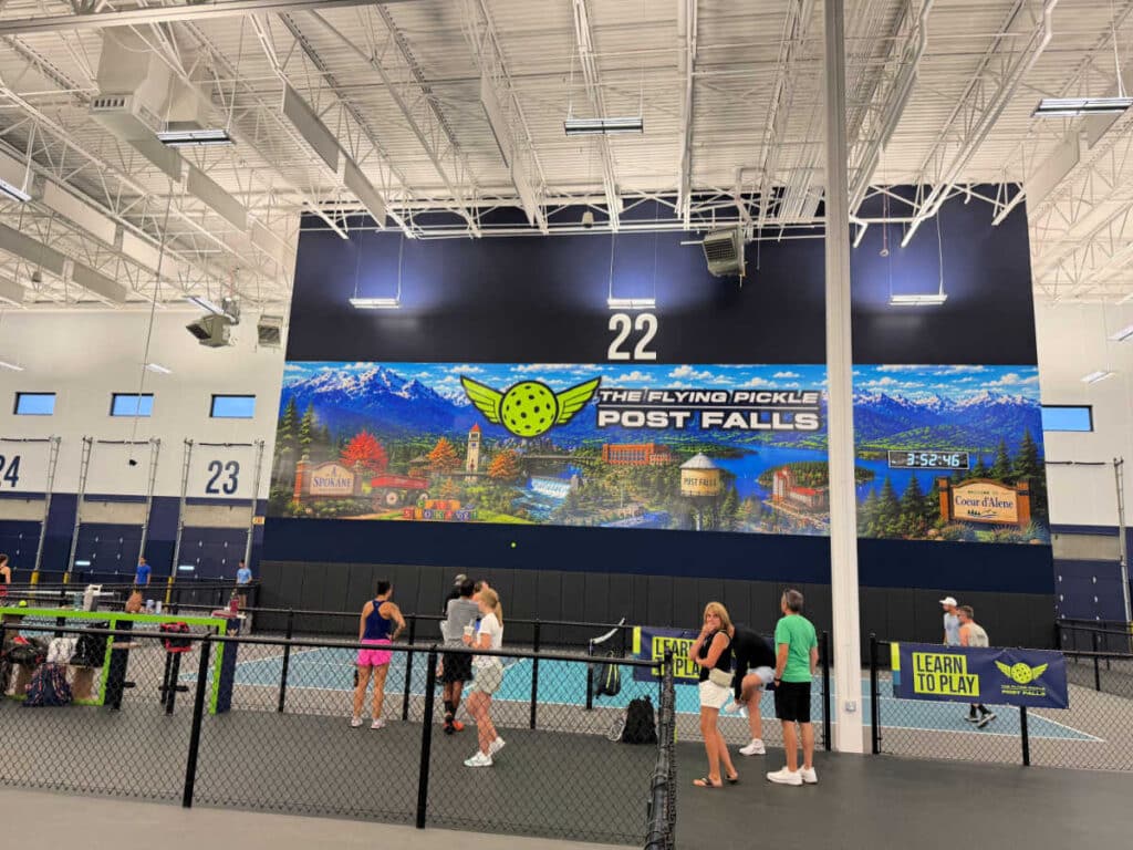 People stand near indoor pickleball courts at The Flying Pickle Post Falls, with a large mural of mountains and the venue name on the back wall. The court numbers 22 and 23 are visible.