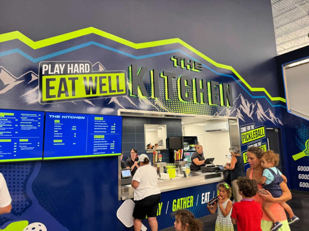 A group of people, including children, stand at a colorful counter labeled The Kitchen, ordering food at a sports venue. The wall says Play Hard Eat Well above the menu boards, capturing the lively spirit of The Flying Pickle Post Falls.