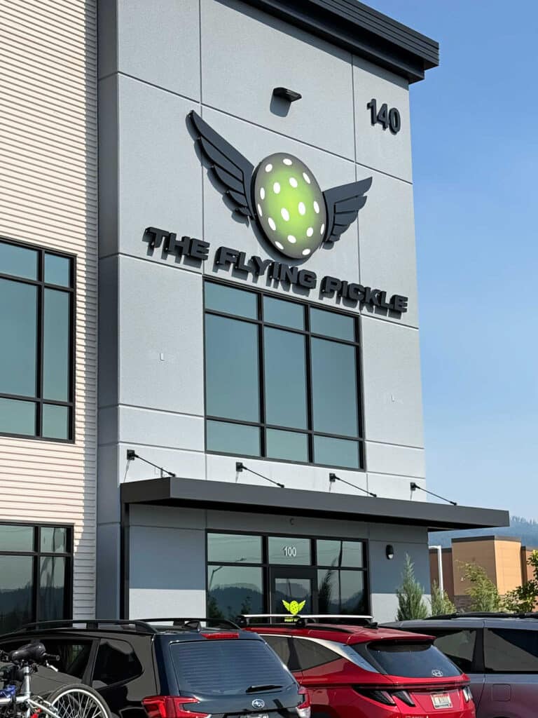 A building with a sign reading The Flying Pickle and a logo of a green pickleball with wings stands prominently in Post Falls. Several cars are parked in front of The Flying Pickle Post Falls, adding to the lively atmosphere.