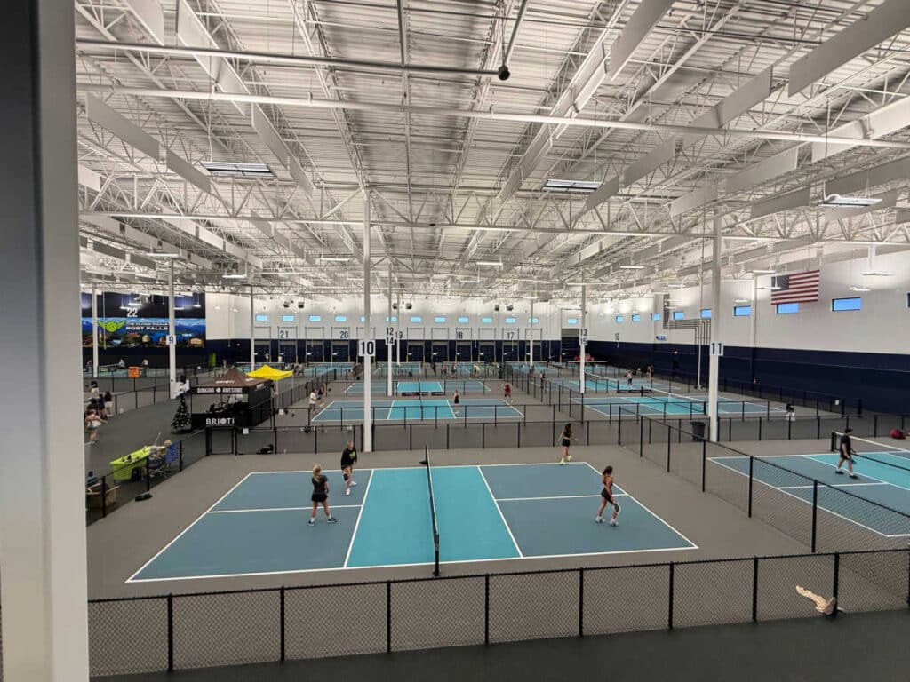 Large indoor sports facility, The Flying Pickle Post Falls, featuring multiple blue pickleball courts, high ceilings, bright lighting, and several people playing. An American flag is displayed on the wall to the right.